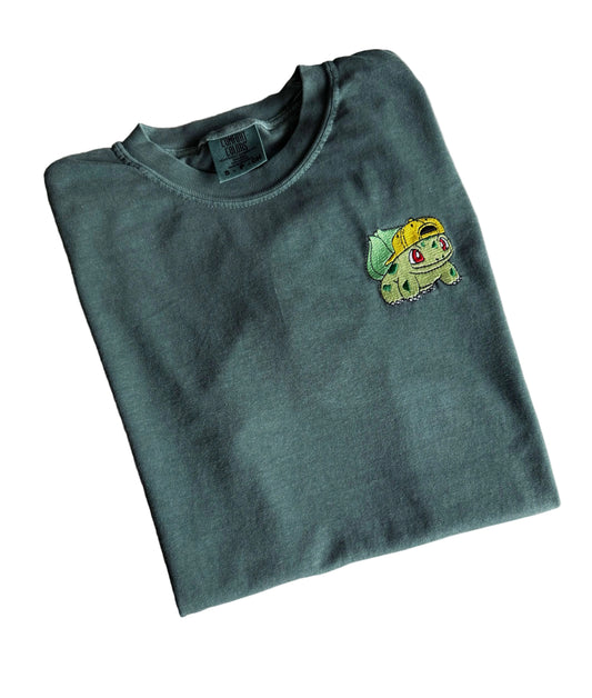 A folded slate blue-gray t-shirt featuring a small embroidered Bulbasaur character on the left chest, laid flat on a light marble-patterned surface.
