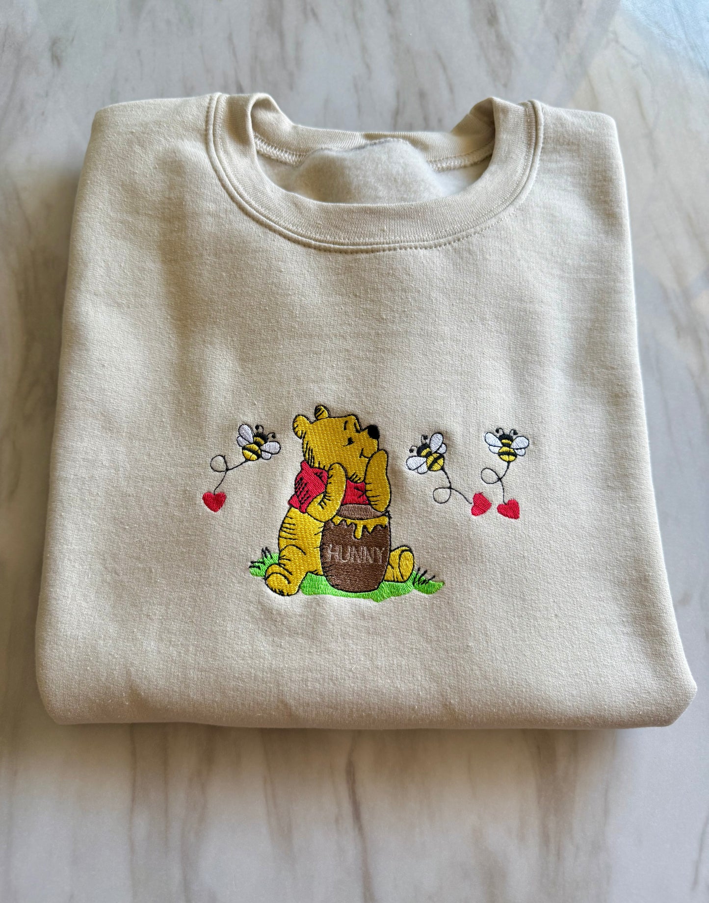 Folded beige sweatshirt with embroidered design of a bear and bees on a marble surface