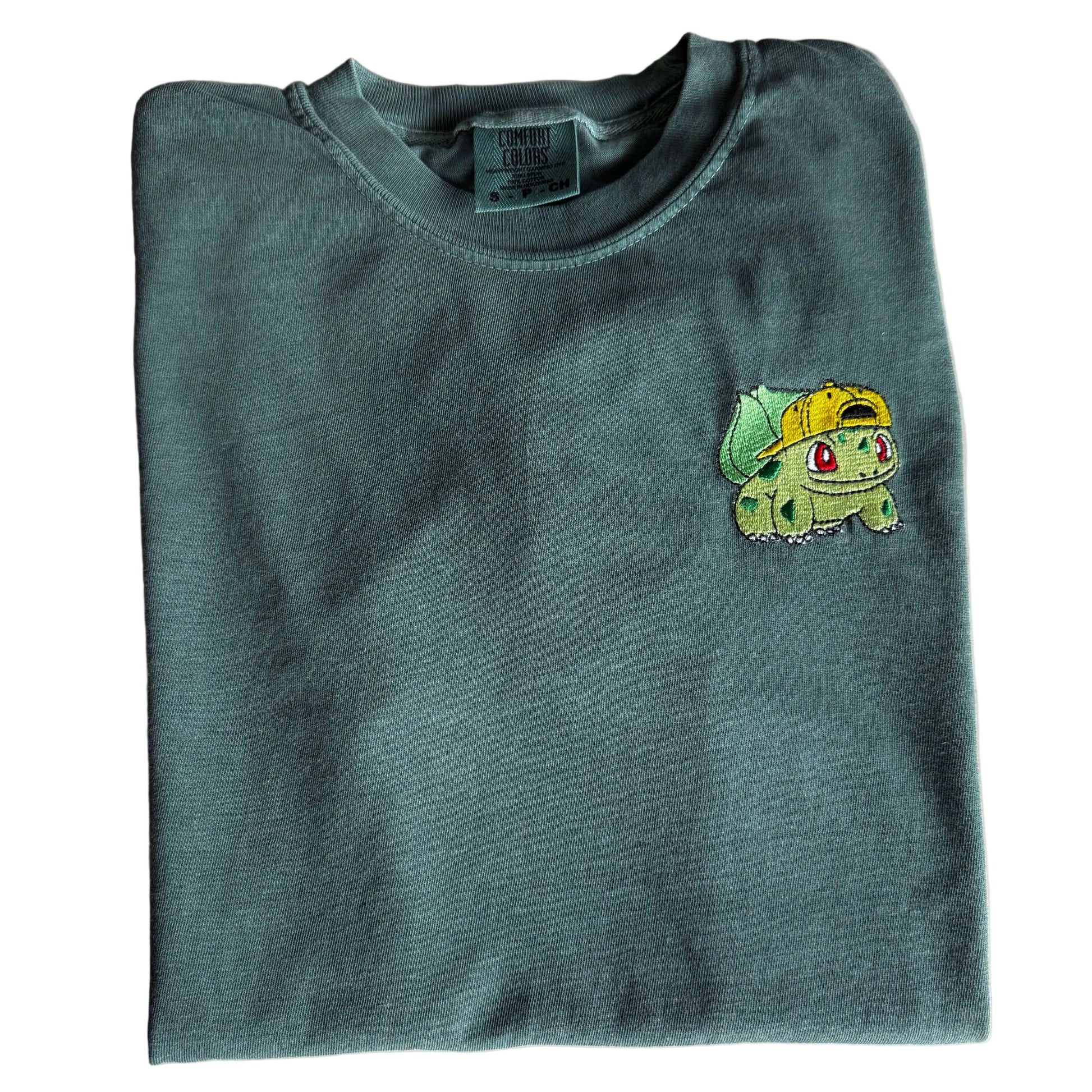 A folded dark green crewneck sweatshirt featuring a small embroidered Bulbasaur character on the left chest, laid flat on a light-colored marble surface.