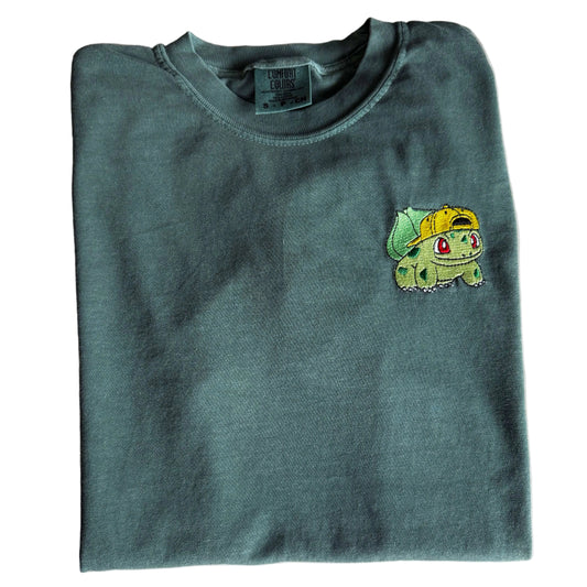 A folded dark green crewneck sweatshirt featuring a small embroidered Bulbasaur character on the left chest, laid flat on a light-colored marble surface.