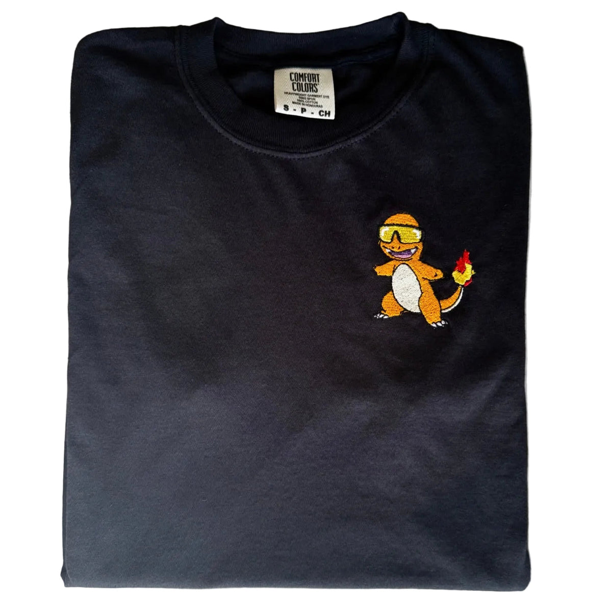 A folded black t-shirt featuring a small embroidered Charmander graphic on the left chest, laid flat on a white marble surface.
