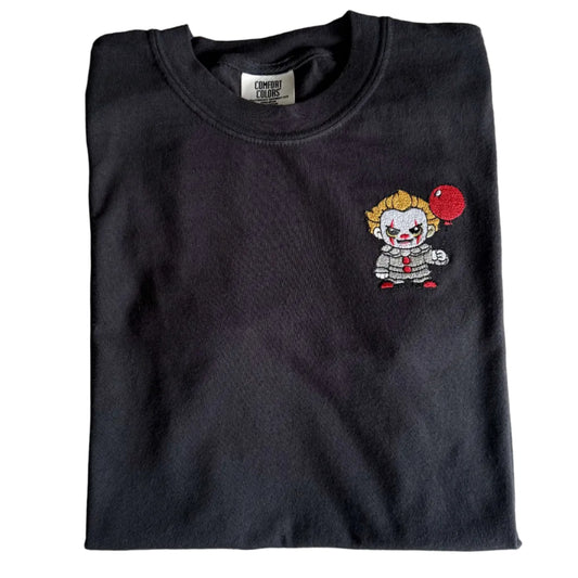 Angled view of a folded black Comfort Colors T-shirt with a small embroidered clown holding a red balloon.